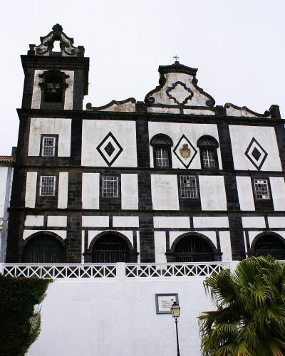 The image shows a church facade with distinctive black basalt stone framing and white plaster infill, featuring diamond-shaped motifs, arched ground-level openings, and a bell tower, situated in a coastal setting with palm trees and a white retain