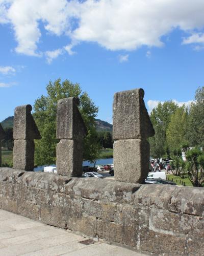 A close-up view of the granite parapet and distinctive merlons of a stone bridge, with a river, trees, and a hillside visible in the background under a blue sky.