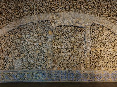 An interior view of a chapel wall and archway constructed entirely from human skulls and bones, featuring a decorative border of blue and yellow patterned tiles at the base.