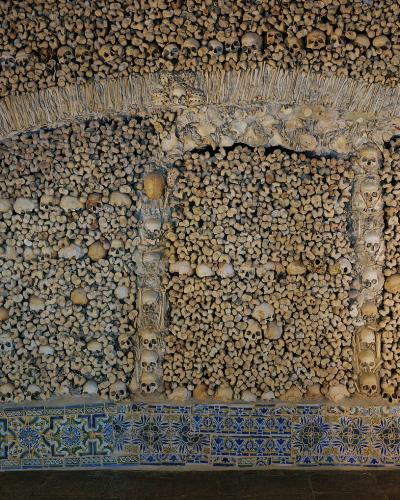 An interior view of a chapel wall and archway constructed entirely from human skulls and bones, featuring a decorative border of blue and yellow patterned tiles at the base.