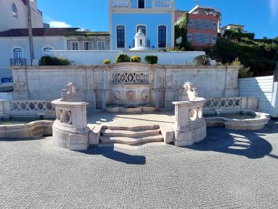 A stone fountain with three spouts (two side basins and a central wall fountain) situated on a cobblestone plaza, with a light blue building featuring a distinctive dark roof and white balustrade in the background.