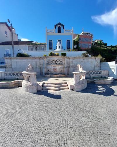 A stone fountain with three spouts (two side basins and a central wall fountain) situated on a cobblestone plaza, with a light blue building featuring a distinctive dark roof and white balustrade in the background.