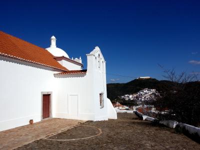 A white-walled hermitage with a red-tiled roof, a small dome, and a bell gable, set against a blue sky with a hillside town and fortress in the background.