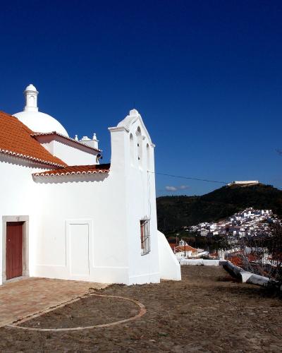 A white-walled hermitage with a red-tiled roof, a small dome, and a bell gable, set against a blue sky with a hillside town and fortress in the background.