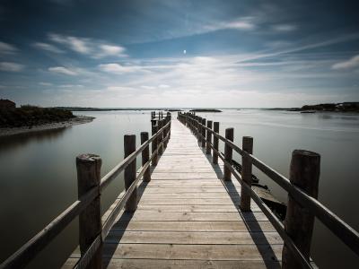 A long wooden boardwalk or pier extends over calm, shallow water towards the horizon, with low-lying vegetation and a building visible on the left bank.