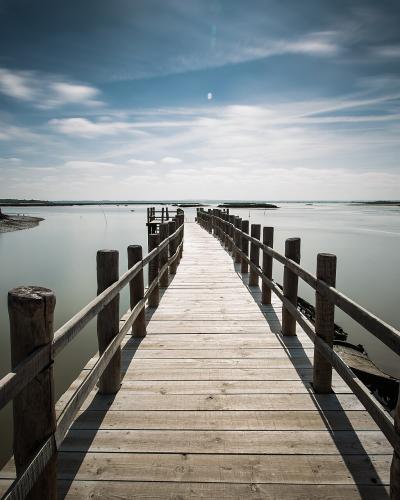 A long wooden boardwalk or pier extends over calm, shallow water towards the horizon, with low-lying vegetation and a building visible on the left bank.
