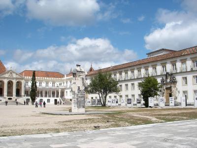 A wide shot of a large, sunlit courtyard surrounded by white university buildings with red-tiled roofs, featuring a central stone monument and an exhibition of framed photos.