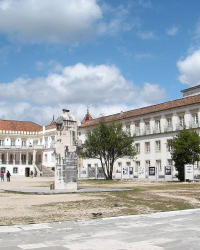 A wide shot of a large, sunlit courtyard surrounded by white university buildings with red-tiled roofs, featuring a central stone monument and an exhibition of framed photos.