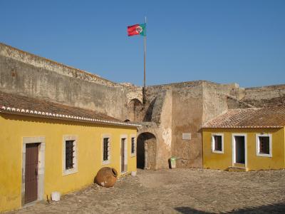 A courtyard within a historic stone fortress featuring yellow buildings with white trim, a large terracotta jar, and a Portuguese flag flying on a tall pole against a clear blue sky.