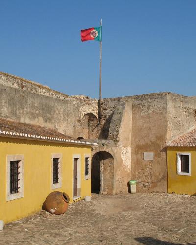 A courtyard within a historic stone fortress featuring yellow buildings with white trim, a large terracotta jar, and a Portuguese flag flying on a tall pole against a clear blue sky.
