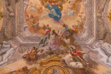 A Baroque-style frescoed ceiling with mythological figures, cherubs, and a central medallion containing a bust portrait, framed by ornate stucco work.