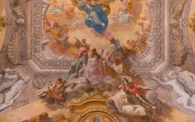 A Baroque-style frescoed ceiling with mythological figures, cherubs, and a central medallion containing a bust portrait, framed by ornate stucco work.