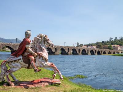 A large, colorful statue of a Roman soldier on horseback positioned on a grassy riverbank with a long, multi-arched Roman stone bridge spanning the river in the background.