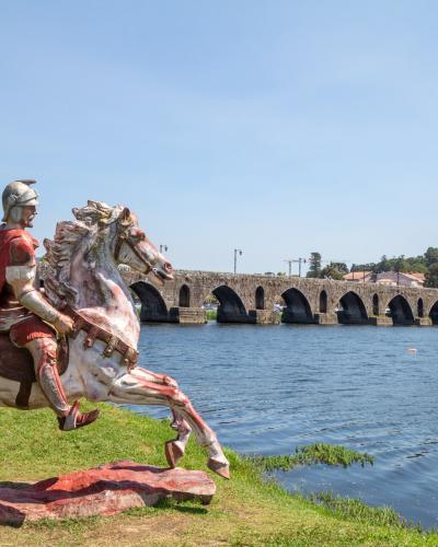 A large, colorful statue of a Roman soldier on horseback positioned on a grassy riverbank with a long, multi-arched Roman stone bridge spanning the river in the background.