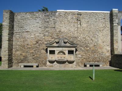 A stone fountain with a decorative pediment and scalloped shell motif set into a high stone wall, flanked by stone benches and a green lawn.