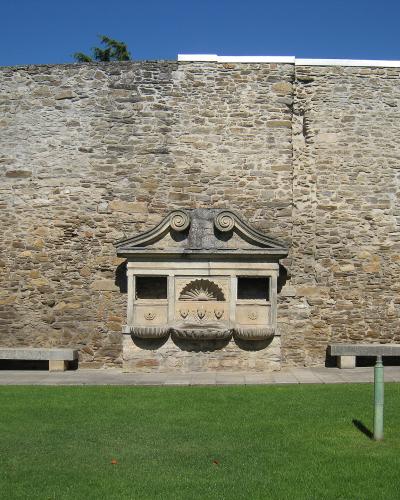 A stone fountain with a decorative pediment and scalloped shell motif set into a high stone wall, flanked by stone benches and a green lawn.