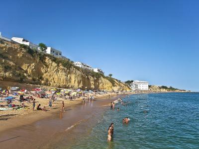 A sunny beach scene featuring golden sand, turquoise water, and numerous people swimming and sunbathing, with a steep, eroded yellow cliff face topped by white buildings rising on the left side.
