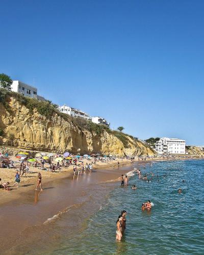 A sunny beach scene featuring golden sand, turquoise water, and numerous people swimming and sunbathing, with a steep, eroded yellow cliff face topped by white buildings rising on the left side.