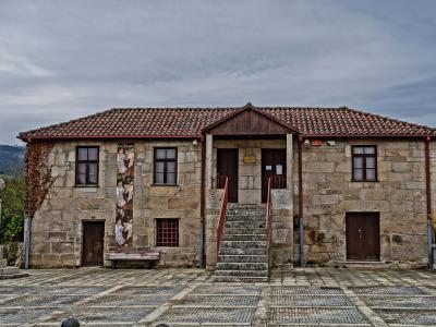 A two-story stone building with a tiled roof, a central staircase leading to double doors, and a stone cross (pelourinho) in the foreground plaza.