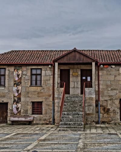 A two-story stone building with a tiled roof, a central staircase leading to double doors, and a stone cross (pelourinho) in the foreground plaza.