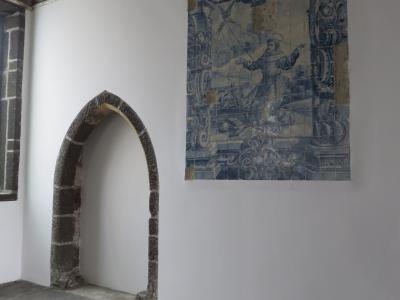 An interior room featuring a stone arched doorway, a wooden plank ceiling, and a large blue and white azulejo tile panel depicting a religious figure on a white wall.