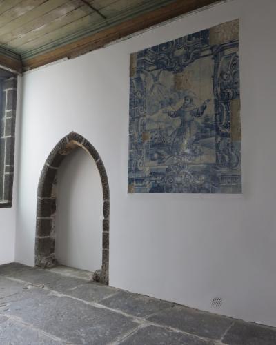 An interior room featuring a stone arched doorway, a wooden plank ceiling, and a large blue and white azulejo tile panel depicting a religious figure on a white wall.