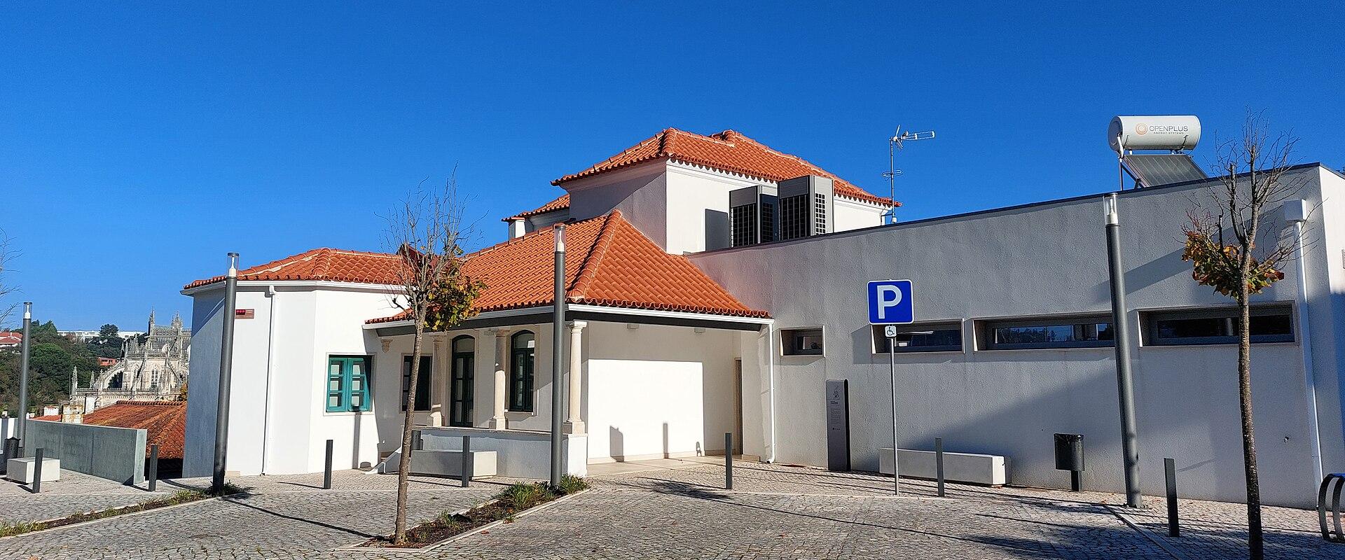A white building with a terracotta tiled roof and a modern grey annex, situated on a cobblestone plaza under a clear blue sky, with a blue parking sign visible.