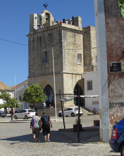 A stone bell tower with a rooster weather vane, situated on a cobblestone street with pedestrians and cars, featuring a street sign reading "RUA DO ARCO".