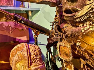 A close-up view of a waxwork automaton figure with a cracked head revealing a brain, positioned next to a telescope and other curiosities under purple lighting.