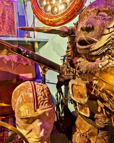 A close-up view of a waxwork automaton figure with a cracked head revealing a brain, positioned next to a telescope and other curiosities under purple lighting.