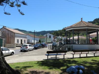 A village square featuring a tiled-roof bandstand (kiosk), benches, parked cars, and traditional white buildings with stone accents under a clear blue sky.