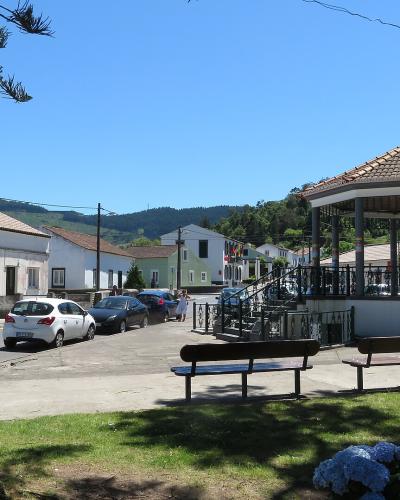 A village square featuring a tiled-roof bandstand (kiosk), benches, parked cars, and traditional white buildings with stone accents under a clear blue sky.