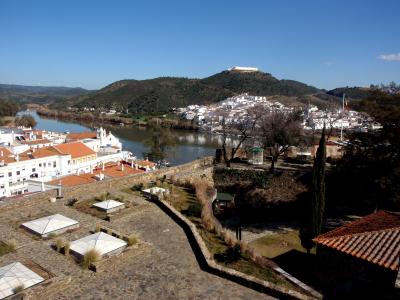 A high-angle view from a stone terrace overlooking the town of Alcoutim, the River Guadiana, and a prominent hill in the distance topped with a large, white-walled fortress structure.