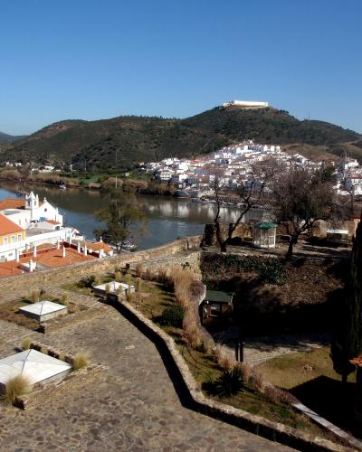 A high-angle view from a stone terrace overlooking the town of Alcoutim, the River Guadiana, and a prominent hill in the distance topped with a large, white-walled fortress structure.