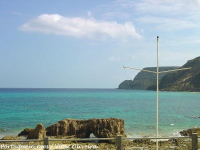 A coastal scene featuring turquoise water, a rocky outcrop with a natural arch in the foreground, and a white cross-shaped structure on a pole to the right, with cliffs in the background.