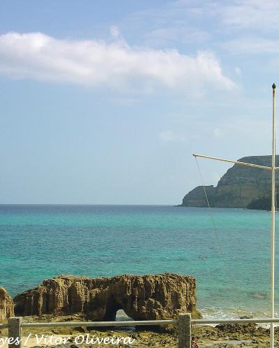 A coastal scene featuring turquoise water, a rocky outcrop with a natural arch in the foreground, and a white cross-shaped structure on a pole to the right, with cliffs in the background.