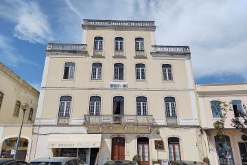A multi-story cream-colored building with arched windows, balconies, and a sign reading "HOSTEL 402" above the ground floor entrance; adjacent storefront includes “retrosaria FILÓ”; parked cars line the street under a partly cloudy sky.