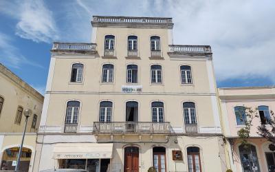 A multi-story cream-colored building with arched windows, balconies, and a sign reading "HOSTEL 402" above the ground floor entrance; adjacent storefront includes “retrosaria FILÓ”; parked cars line the street under a partly cloudy sky.