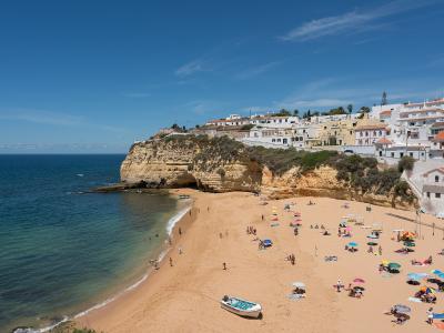 A high-angle view of a sandy beach with golden cliffs and white buildings on the headland, featuring a small boat on the sand and people relaxing under umbrellas.
