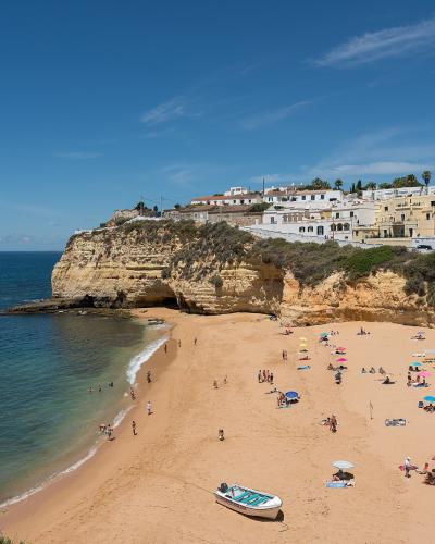 A high-angle view of a sandy beach with golden cliffs and white buildings on the headland, featuring a small boat on the sand and people relaxing under umbrellas.