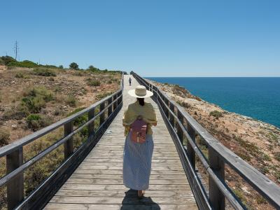 A person wearing a straw hat and long skirt walks away from the camera along a long, straight wooden boardwalk with railings, overlooking the ocean on the right and dry coastal scrub on the left.