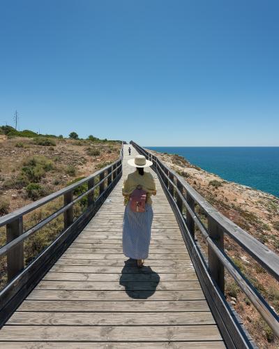 A person wearing a straw hat and long skirt walks away from the camera along a long, straight wooden boardwalk with railings, overlooking the ocean on the right and dry coastal scrub on the left.