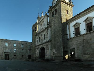 A large stone cathedral with a prominent bell tower and an ornate Baroque-style facade, situated in a paved public square under a clear blue sky.