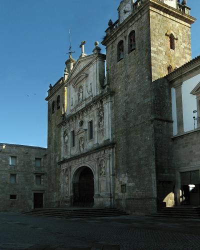 A large stone cathedral with a prominent bell tower and an ornate Baroque-style facade, situated in a paved public square under a clear blue sky.