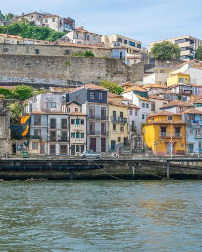 A view from the Douro River looking up at the colorful, terraced buildings of the Ribeira district in Porto, Portugal, with a stone retaining wall and a small boat on the water.