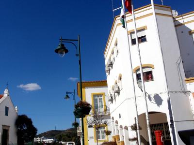A sunlit street scene in a Portuguese town featuring white buildings with yellow trim, a cobblestone road, a green lamppost, and three flagpoles flying the Portuguese national flag, the European Union flag, and a third flag (likely regional or mun