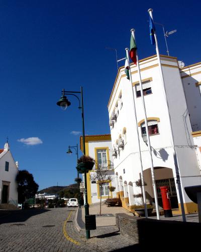 A sunlit street scene in a Portuguese town featuring white buildings with yellow trim, a cobblestone road, a green lamppost, and three flagpoles flying the Portuguese national flag, the European Union flag, and a third flag (likely regional or mun