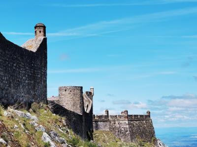 A stone fortress with crenellated walls and towers situated on a steep, rocky hillside under a blue sky.