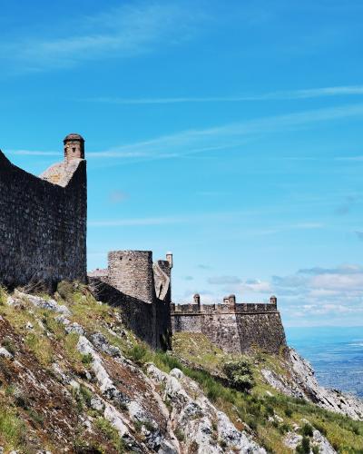 A stone fortress with crenellated walls and towers situated on a steep, rocky hillside under a blue sky.