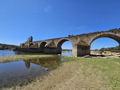 A long, multi-arched medieval stone bridge in a state of partial ruin, spanning a river with low water levels under a clear blue sky.